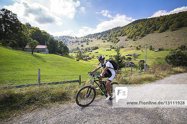 Male athlete cycling on dirt road by field against sky