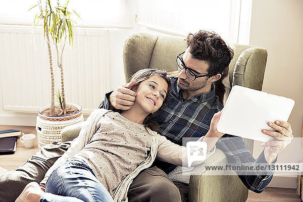 Vater sieht Tochter beim Lesen eines digitalen Tabletts an  während er auf einem Stuhl sitzt