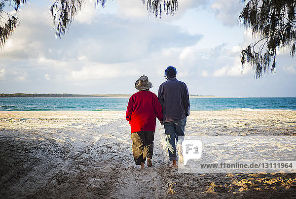 Rear view of senior couple holding hands while walking on sandy beach