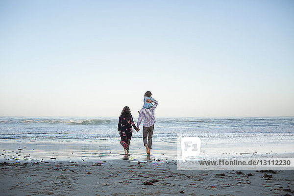 Rear view of man piggybacking daughter while holding woman's hand on shore at beach against clear sky