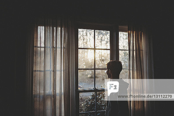 Seitenansicht eines Teenagers  der durch ein Fenster schaut  während er zu Hause steht