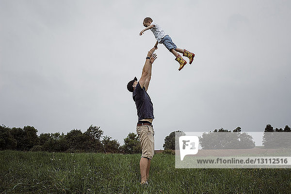 Glücklicher Vater wirft Sohn in die Luft  während er auf Grasfeld gegen den Himmel im Park steht