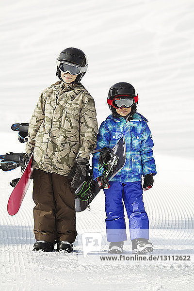 Smiling siblings carrying snowboards while standing on snow covered field