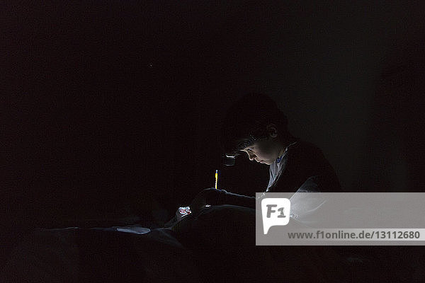 Seitenansicht eines Jungen mit beleuchteter Stirnlampe  der Hausaufgaben schreibt  während er zu Hause in der Dunkelkammer auf dem Bett sitzt