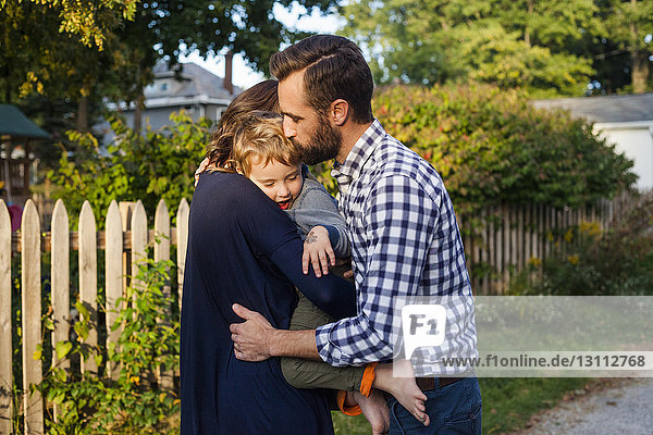 Seitenansicht von Eltern  die den Sohn umarmen und küssen  während sie im Garten vor Pflanzen stehen