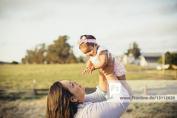 Mother picking up daughter while standing at ranch against sky during sunset