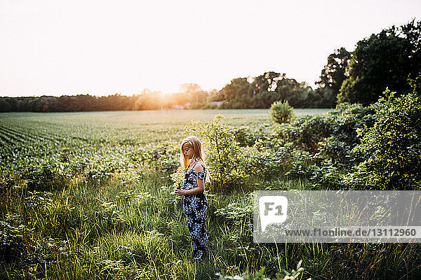 Side view of girl holding flowers while standing on grassy field against clear sky during sunset
