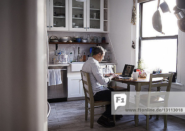 Side view of man using laptop computer while sitting at dining table in kitchen