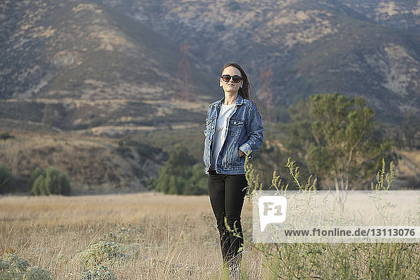 Selbstbewusste junge Frau mit Händen in den Taschen  die auf dem Feld gegen den Berg steht