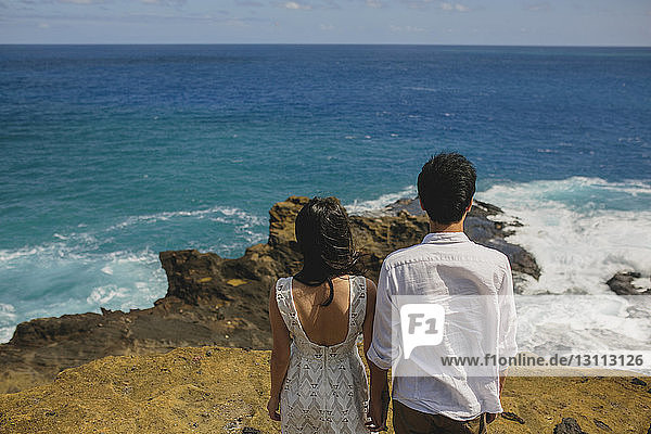 Rückansicht eines Paares  das sich an den Händen hält und am Strand gegen den Himmel steht