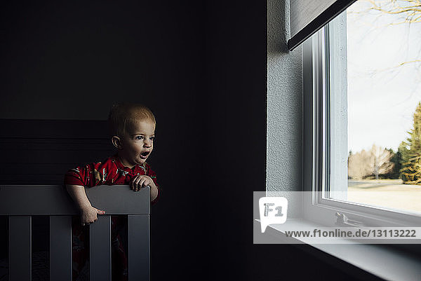 Kleiner Junge mit offenem Mund  der durch ein Fenster schaut  während er zu Hause am Stuhl steht