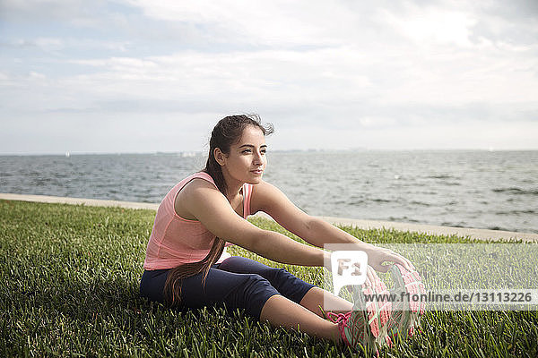 Young woman touching toes while sitting on grassy field against sea