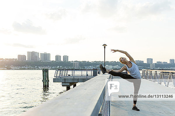 Frau in voller Länge streckt ihr Bein auf dem Geländer an der Brücke gegen den Himmel in der Stadt