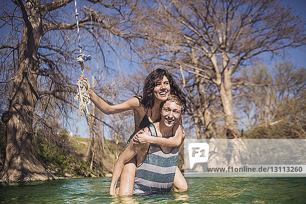 Porträt einer fröhlichen Freundin  die an einem sonnigen Tag ihrem Freund im Fluss vor kahlen Bäumen Huckepack gibt