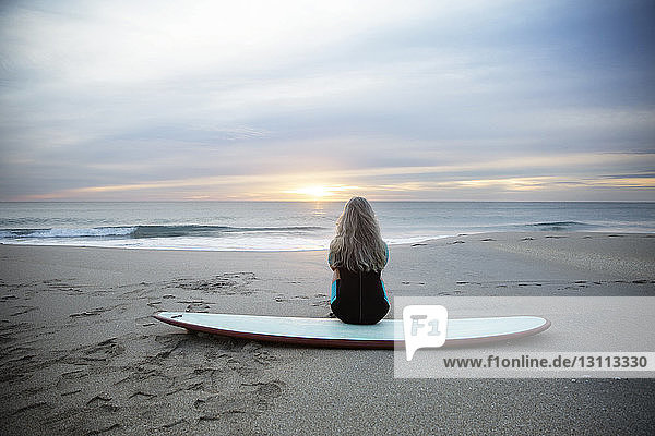 Rückansicht einer Surferin  die bei Sonnenuntergang am Strand von Delray auf einem Surfbrett sitzt