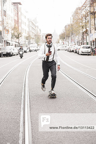 Businessman holding phone while skateboarding on road in city