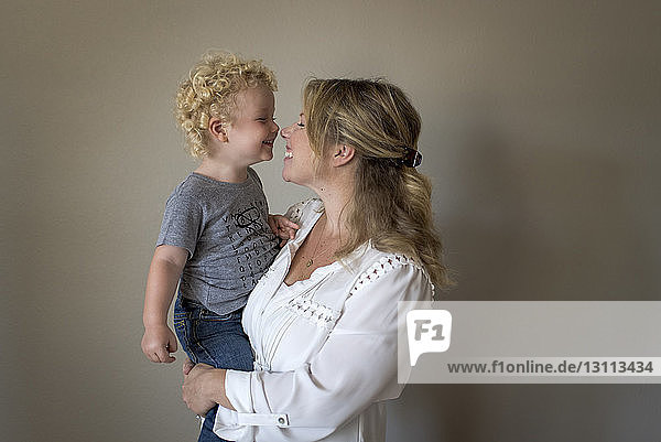 Cheerful mother carrying son against wall at home