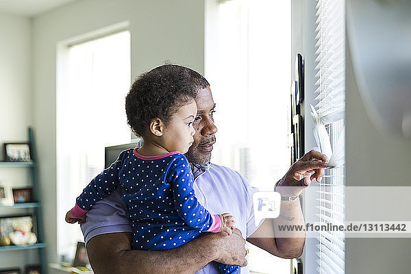 Father and daughter looking through window blinds at home