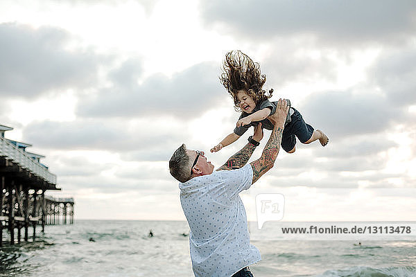 Playful father throwing son in air while playing against cloudy sky at beach