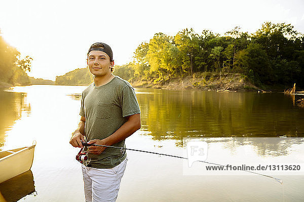 Portrait of young man with fishing rod standing at lakeshore against clear sky during sunset
