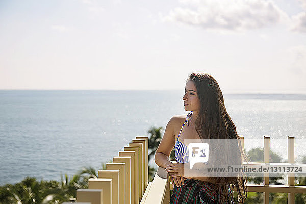 Thoughtful woman standing by railing at balcony against sea on sunny day