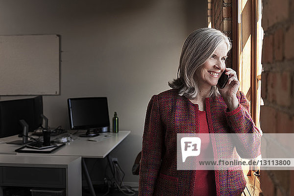 Glückliche Geschäftsfrau beantwortet Smartphone  während sie im Kreativbüro durchs Fenster schaut