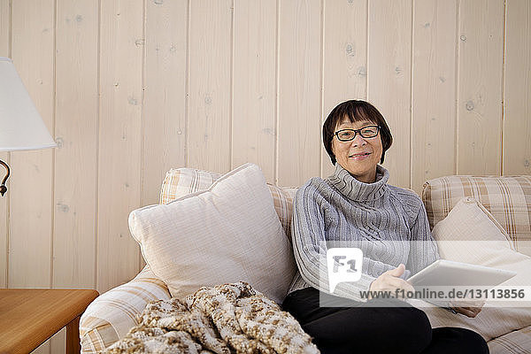 Porträt einer glücklichen älteren Frau  die einen Tablet-Computer hält  während sie zu Hause auf dem Sofa sitzt