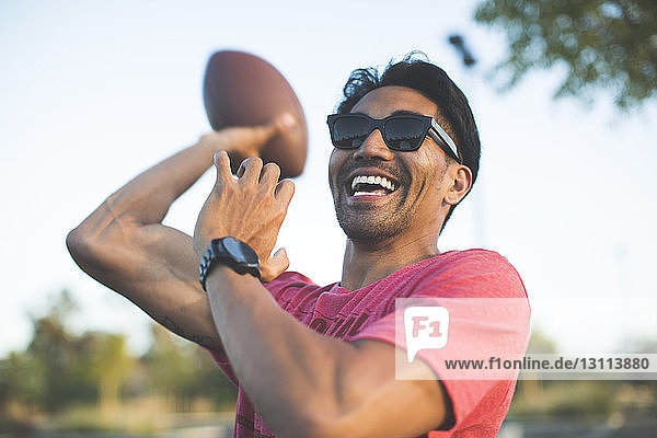 Cheerful man in sunglasses playing with American football against sky