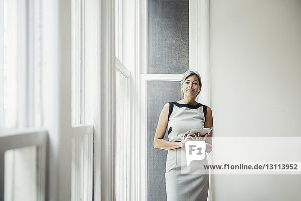 Porträt einer selbstbewussten Geschäftsfrau  die einen Tablet-Computer hält  während sie in einem kreativen Büro am Fenster steht