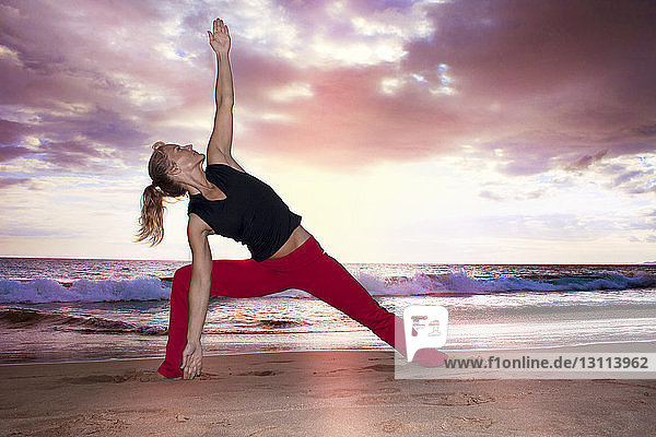 Woman practicing yoga on shore against cloudy sky during sunset