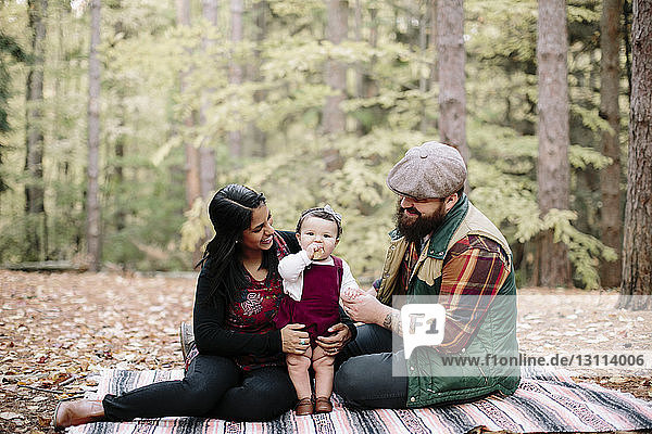 Happy parents looking at daughter eating while relaxing on blanket in forest
