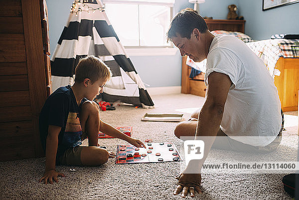 Vater und Sohn spielen Dame  während sie zu Hause auf einem Teppich sitzen