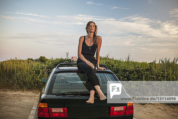 Nachdenkliche Frau sitzt auf Autodach auf Feld gegen Himmel