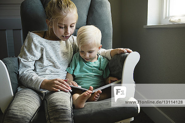 Schwester und Bruder schauen sich ein Buch an  während sie zu Hause auf einem Stuhl sitzen