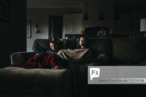 Brothers under blankets watching TV while sitting on sofa at home in darkroom