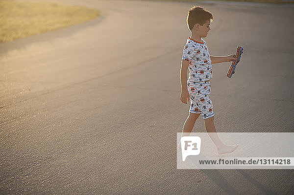 Seitenansicht eines Jungen  der eine Spielzeugeisenbahn hält  während er bei Sonnenuntergang auf der Straße geht