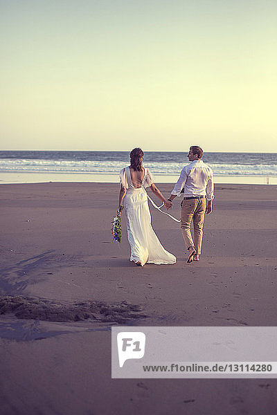 Rückansicht eines frisch vermählten Paares  das sich bei Sonnenuntergang am Strand bei klarem Himmel an den Händen hält