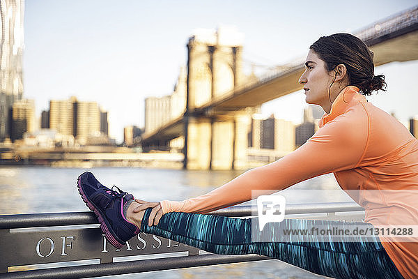 Nachdenkliche Athletin beim Beinstrecken auf der Promenade mit der Brooklyn Bridge im Hintergrund