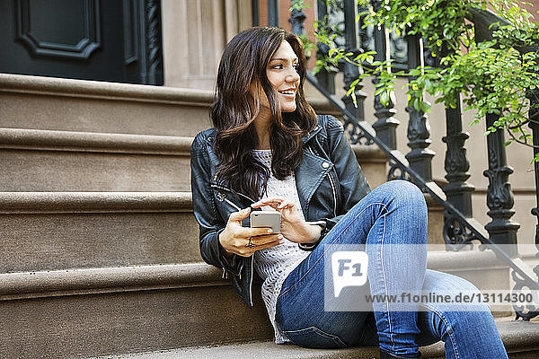 Nachdenkliche Frau hält Smartphone in der Hand  während sie auf Stufen sitzt