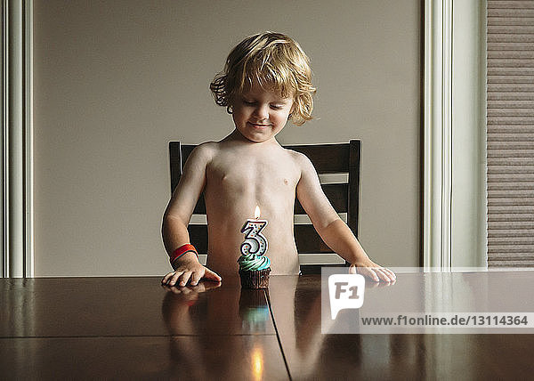 Junge ohne Hemd schaut Geburtstagskuchen an  während er an einem Holztisch steht