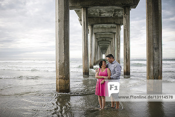 Lächelndes Paar steht am Ufer unter einer Brücke am Strand
