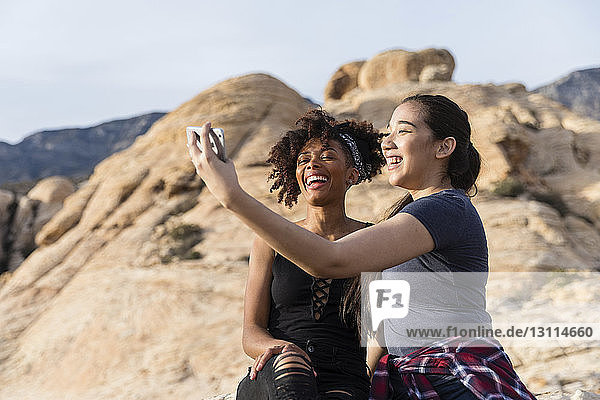 Female friends taking selfie while sitting by rock formation against clear sky