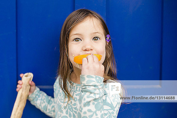 Porträt eines süßen Mädchens  das orange vor blauer Tür isst