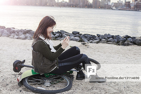 Seitenansicht einer glücklichen Frau  die ein Smartphone benutzt  während sie am Strand auf dem Fahrrad sitzt