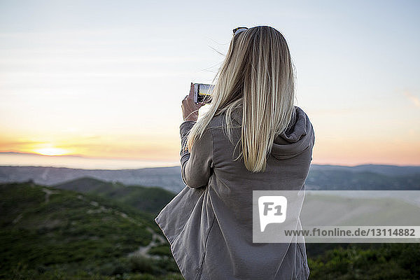 Rear view of woman photographing with mobile phone while standing on mountain against sky