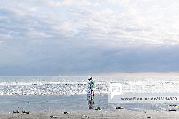 Loving couple standing on shore at beach against cloudy sky