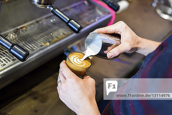 Mittelteil eines Barista  der Milch in Kaffee gießt  während er im Café ein schaumiges Getränk zubereitet