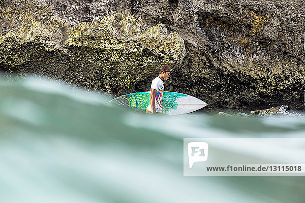 Seitenansicht eines Mannes mit Surfbrett  der an einer Felsformation im Meer steht