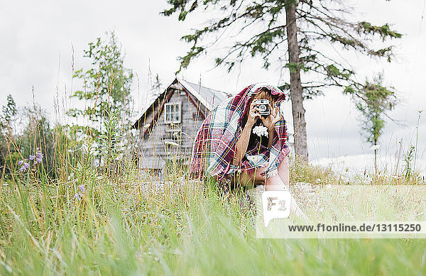 In eine Decke gewickelte Frau fotografiert mit Sofortbildkamera  während sie auf einem Grasfeld sitzt
