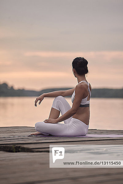 Side view of woman practicing yoga while sitting on pier by lake against cloudy sky during sunset